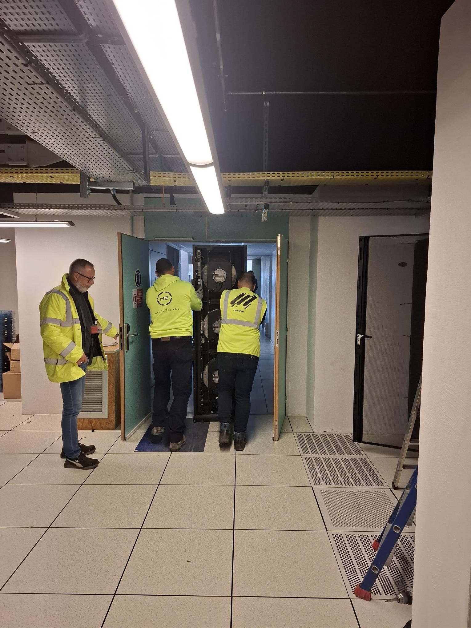 Trois techniciens en uniforme jaunes travaillent sur des équipements dans un serveur.